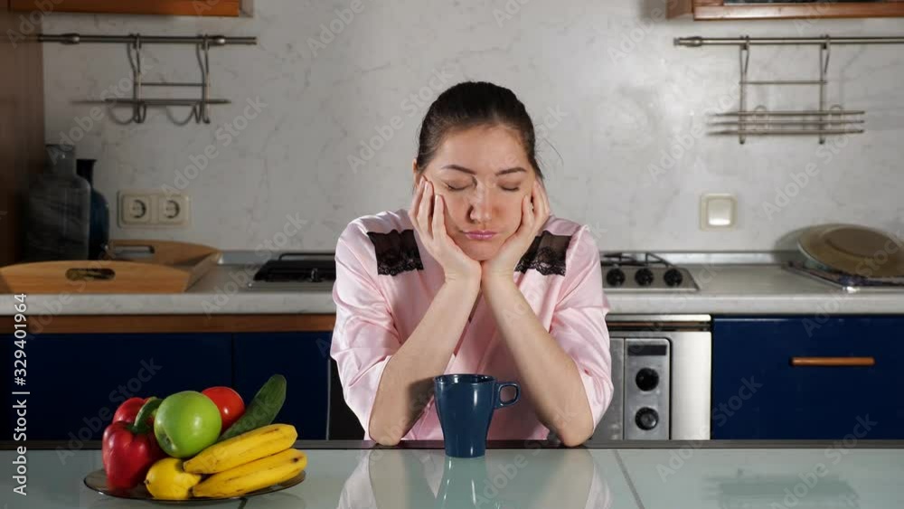 upset sleepy girl in pink home bathrobe sits at kitchen table with cup of coffee and falls asleep closeup upset sleepy girl in pink home bathrobe sits at kitchen table with cup of coffee and falls asleep closeup