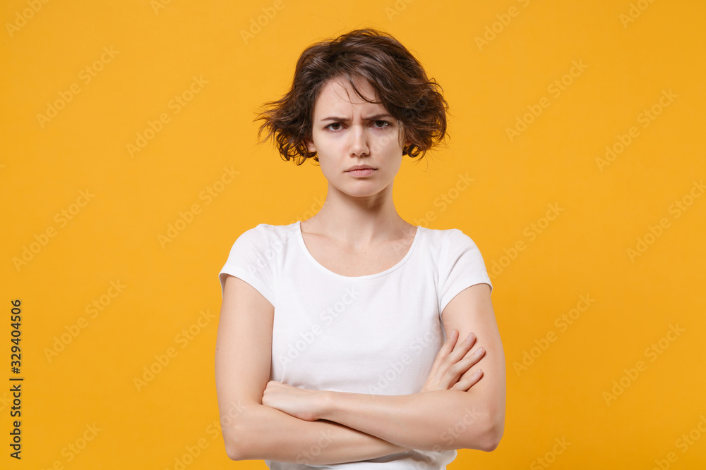 Dissatisfied young brunette woman in white t-shirt posing isolated on yellow orange background studio portrait. People sincere emotions lifestyle concept. Mock up copy space. Holding hands crossed. Dissatisfied young brunette woman in white t-shirt posing isolated on yellow orange background studio portrait. People sincere emotions lifestyle concept. Mock up copy space. Holding hands crossed.