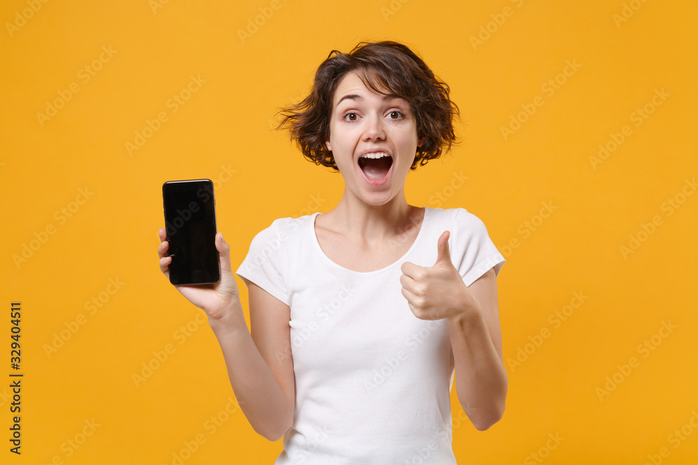 Shocked young brunette woman in white t-shirt posing isolated on yellow orange background. People lifestyle concept. Mock up copy space. Hold mobile phone with blank empty screen, showing thumb up. Shocked young brunette woman in white t-shirt posing isolated on yellow orange background. People lifestyle concept. Mock up copy space. Hold mobile phone with blank empty screen, showing thumb up.