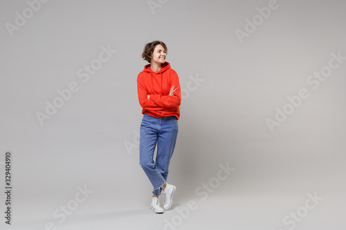 Funny young brunette woman girl in casual red hoodie, blue jeans posing isolated on grey background studio portrait. People lifestyle concept. Mock up copy space. Holding hands crossed, looking aside.