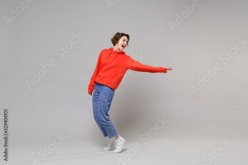 Side view of cheerful young brunette woman girl in casual red hoodie blue jeans posing isolated on grey background. People lifestyle concept. Mock up copy space. Stand on toes looking aside, dancing.