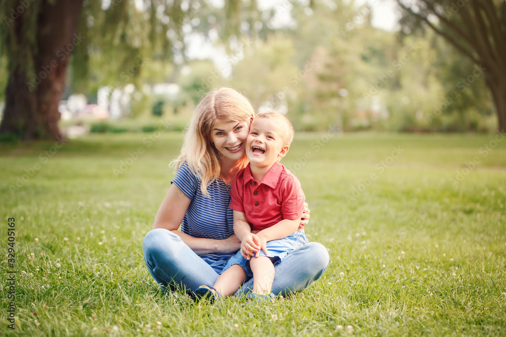Young smiling Caucasian mother and laughing boy toddler son sitting on grass in park. Family mom and child hugging having fun outdoor on a summer day. Happy authentic family childhood lifestyle. Young smiling Caucasian mother and laughing boy toddler son sitting on grass in park. Family mom and child hugging having fun outdoor on a summer day. Happy authentic family childhood lifestyle.