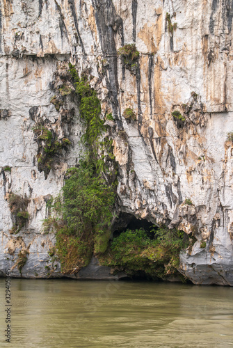 Guilin, China - May 10, 2010: Along Li River. Green foliage covers cave opening in white, black and brown vertical cliff, on greenish water level.