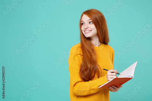 Smiling young redhead woman girl in yellow knitted sweater posing isolated on blue turquoise background in studio. People lifestyle concept. Mock up copy space. Write notes in notebook, looking aside.