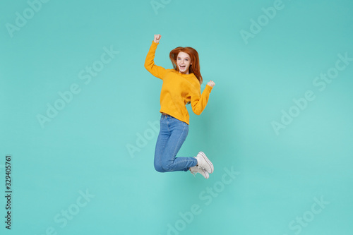 Cheerful young redhead woman girl in yellow knitted sweater posing isolated on blue turquoise background studio portrait. People lifestyle concept. Mock up copy space. Jumping doing winner gesture.