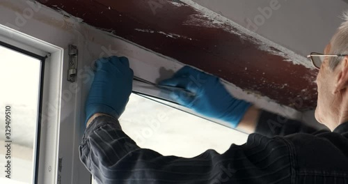 Professional repairman in blue gloves and a gray shirt carefully removes the plastic seal from the white window with a tool. repair in a new apartment. close-up camera movement
