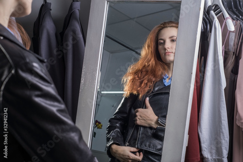 portrait of a beautiful red-haired woman thirty-seven years old, in a women's clothing store, a girl tries on a leather jacket made of high-quality fabric by the mirror