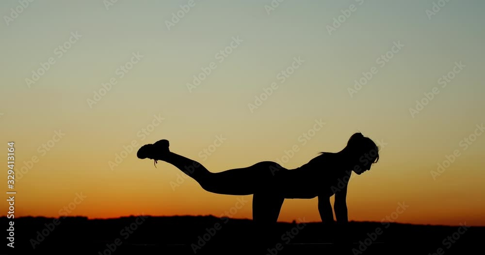 Young woman exercising working out silhouetted against a colorful orange sunset on a wooden deck doing leg raises and stretched to strengthen her muscles