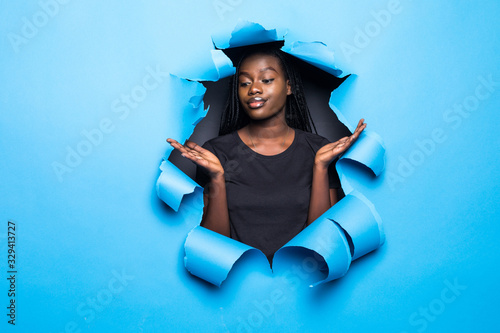 Young confused african woman with raised hands posing from blue paper hole
