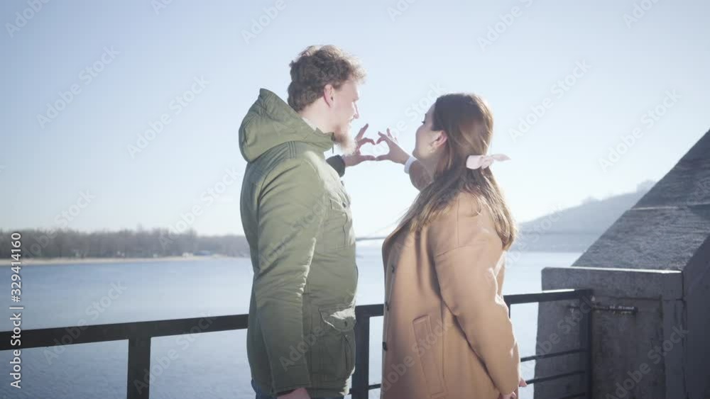 Happy young Caucasian man and woman making heart shape with hands at the background of blue autumn sky. Smiling couple looking at each other with love as standing on river bank outdoors. Love, romance Happy young Caucasian man and woman making heart shape with hands at the background of blue autumn sky. Smiling couple looking at each other with love as standing on river bank outdoors. Love, romance