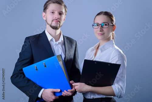 Portrait of young people, businessmen smiling, stand in business suits, holding office folders for papers in their hands. Studio photo on a gray background