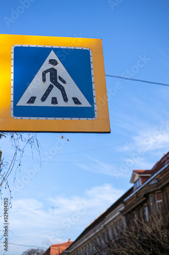 Metal blue roadsign with yellow edging which means - pedestrian crossing. Blue sky, the road in town and a house.