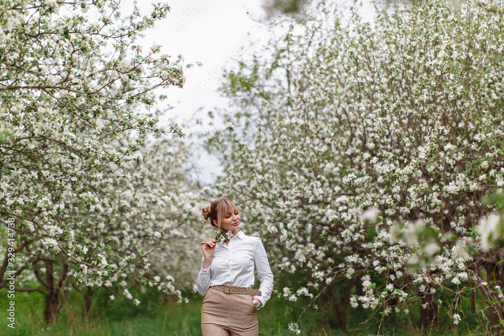 Beautiful young blonde woman in white shirt posing under apple tree in blossom in Spring garden Beautiful young blonde woman in white shirt posing under apple tree in blossom in Spring garden