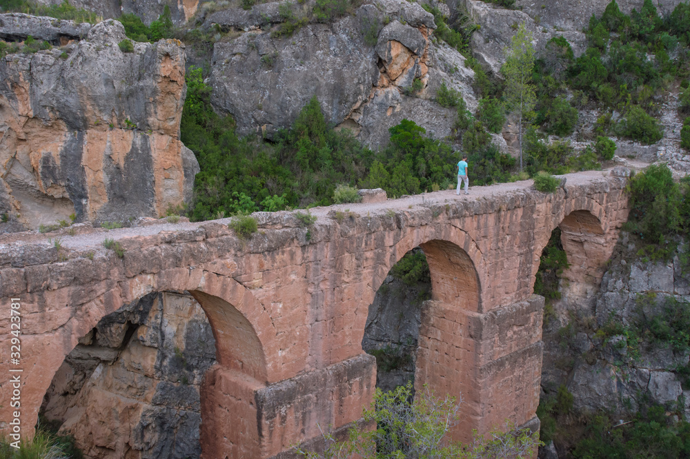 Ancient roman aqueduct with a man walking on it surrounded by rocky mountains in Chelva, valencian community, Spain