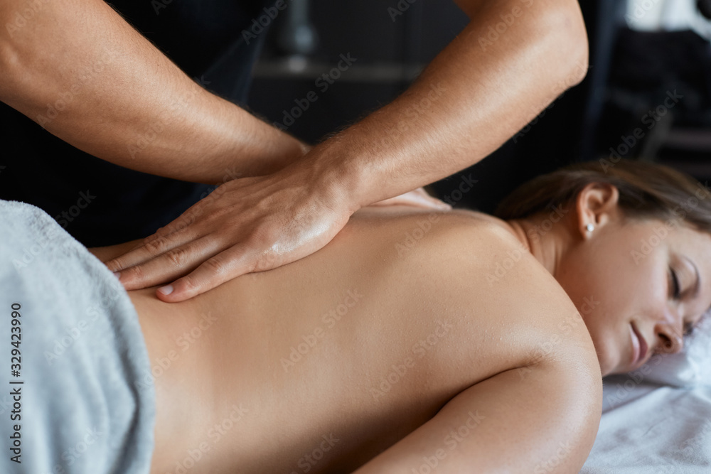 Fototapeta premium Young beautiful woman enjoying back and shouders massage in spa.Professional massage therapist is treating a female patient in apartment.Relaxation,beauty,body and face treatment concept.Home massage.