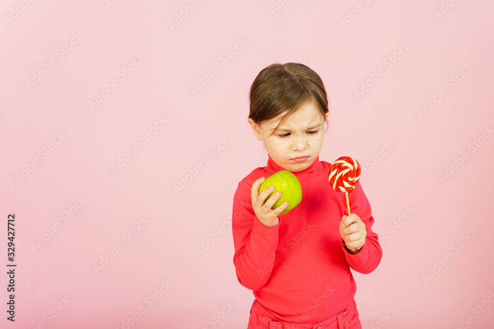 Little girl chooses between a lollipop and a green apple. The concept of proper nutrition. A child in a pink studio holds a sweet of sugar in his hand and an apple. Difficulty of choice Little girl chooses between a lollipop and a green apple. The concept of proper nutrition. A child in a pink studio holds a sweet of sugar in his hand and an apple. Difficulty of choice