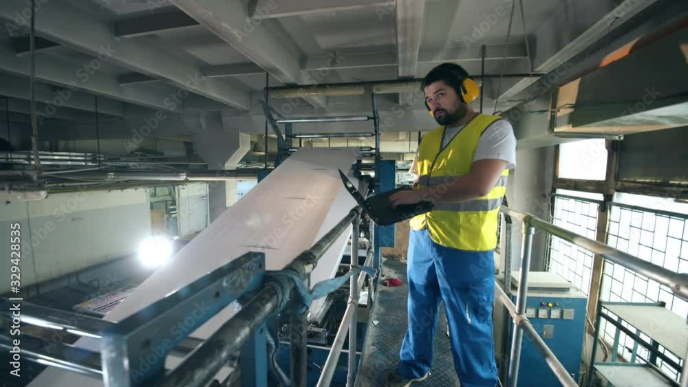 Male worker with a laptop is observing paper-producing machine. Engineer, worker at a factory facility.