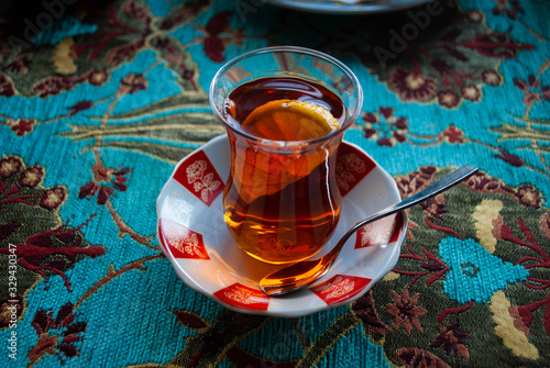 Armud tea glass on a saucer with a small spoon on a blue decorative tablecloth with animals. The glass in the shape of a pear used in Azerbaijan is also known as armud glass. Tea with lemon photo