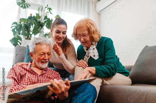 Older man is sitting on the floor in living room and holding family photo album. Happy family evokes happy memories by spending quality time together.