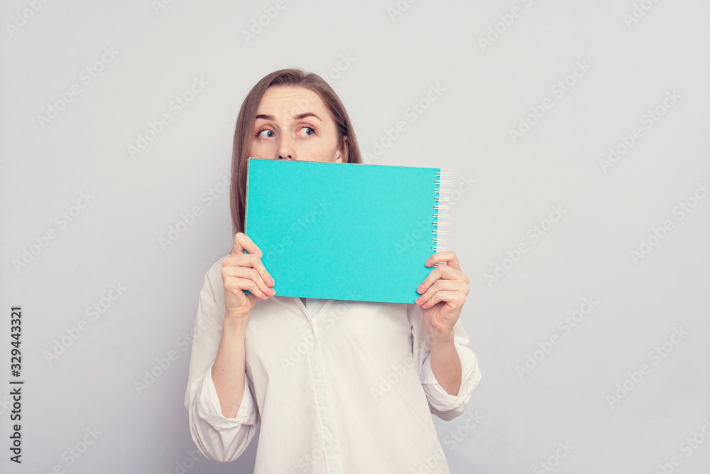 Scared student girl hides behind a notebook, grey background, copy space Scared student girl hides behind a notebook, grey background, copy space