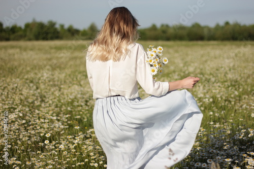 stylish young blonde woman walking in a field with daisies on a sunny summer day.