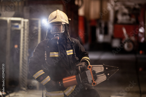 firefighter portrait wearing full equipment, oxygen mask, and power hydraulic cutting tool, smoke and fire trucks in the background.