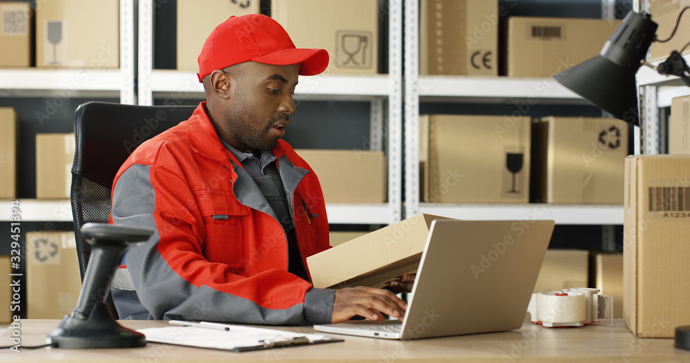 Young African American mailman in red uniform and cap sitting at desk in postal office store and working at laptop computer. Portrait of male courier filling in invoice and entering data. Young African American mailman in red uniform and cap sitting at desk in postal office store and working at laptop computer. Portrait of male courier filling in invoice and entering data.