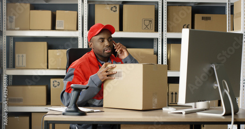 African American man talking on mobile phone while working at post office store and on computer. Postman scanning bar code with scanner, registering box and filling in invoice, speaking on cellphone.