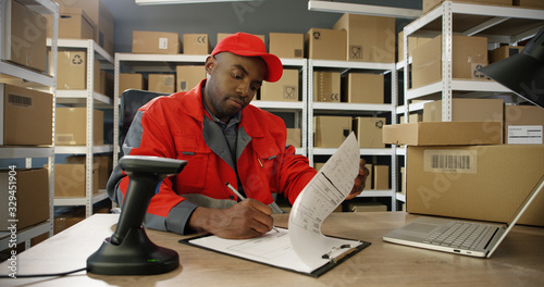 фотографія African American young man in uniform sitting at table and typing on latop computer while registering mail box in post office