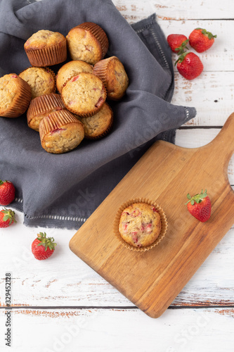 Bowl of Freshly Baked Homemade Strawberry Muffins in a Wood Bowl; One Isolated on Wood Cutting Board in Front; Fresh Strawberrries Scattered Around; White Wood Tabletop photo
