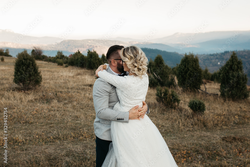 Beautiful couple having a romantic moment on their weeding day, in mountains at sunset. Bride is in a white wedding dress with a bouquet of sunflowers in hand, groom in a suit. Playing together.