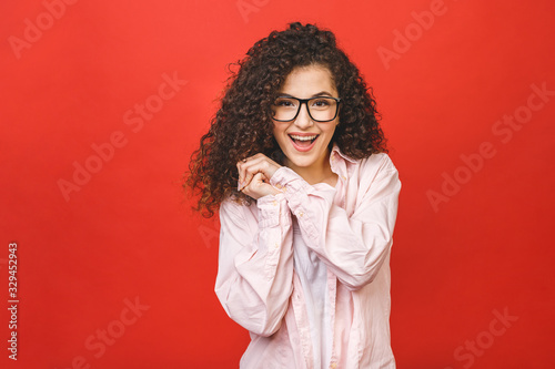 Photo of cheerful beautiful young woman standing isolated over red wall background. Looking camera showing winner gesture. © denis_vermenko