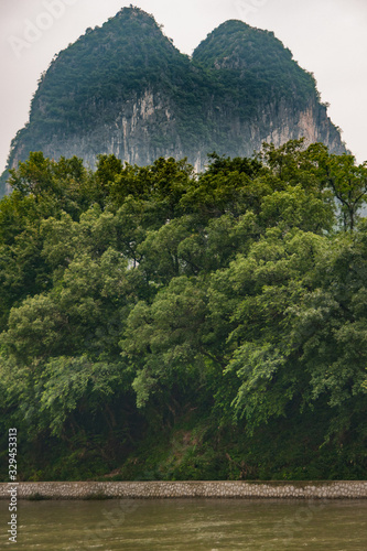 Guilin, China - May 10, 2010: Along Li River. Portrait of twin forested karst mountains almost vertically towering over the forest and greenish water under silver sky. 