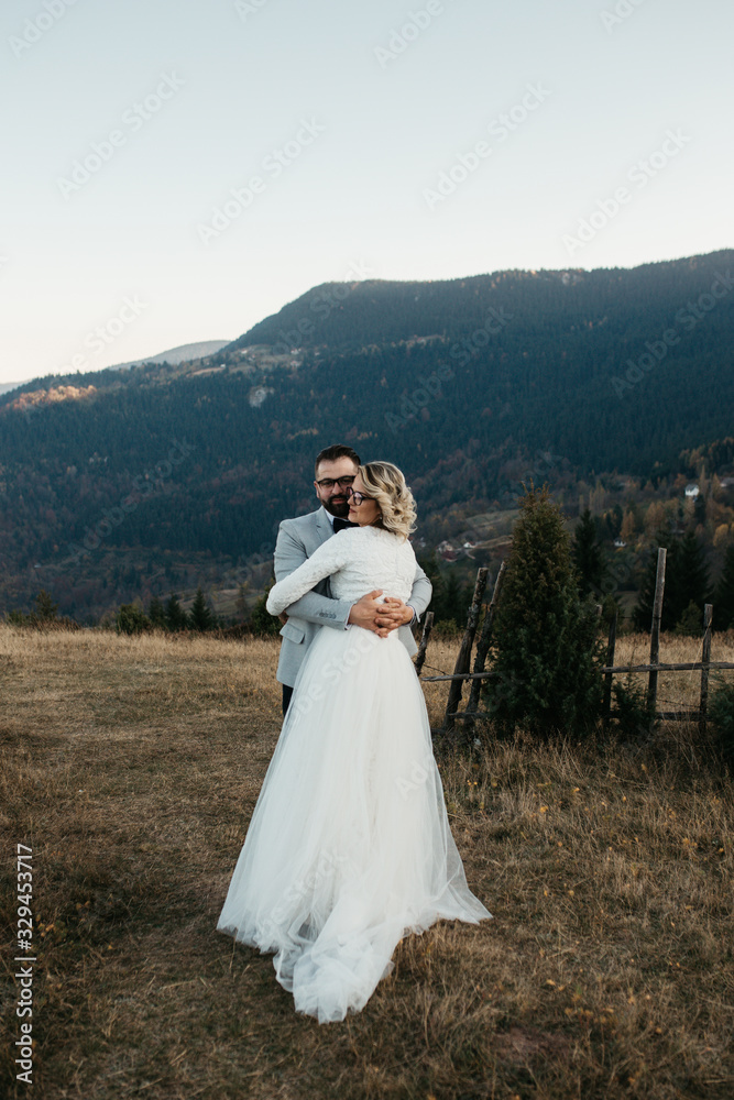 Beautiful couple having a romantic moment on their weeding day, in mountain, sunset.She is in a white wedding dress with a bouquet of sunflowers in hand,groom in a suit. Groom holds bride in his hands