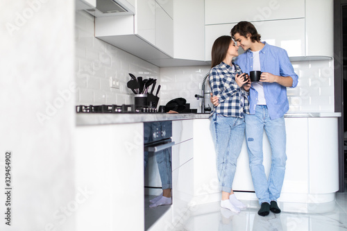 Romantic couple at home. Attractive young woman and handsome man are enjoying spending time together while standing on light modern kitchen with cup of coffee in hands. photo