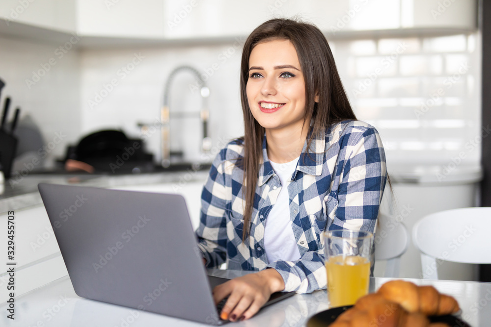 Portrait of pretty woman using laptop at table in kitchen interior while having breakfast at home Portrait of pretty woman using laptop at table in kitchen interior while having breakfast at home