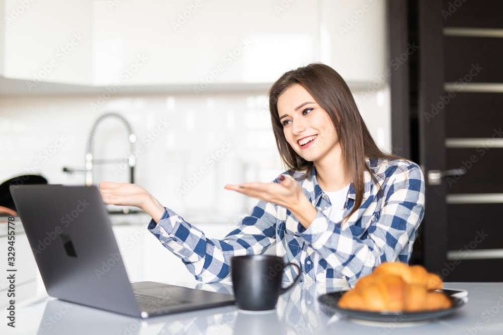 Young pretty woman talking on video call and waving hand while sitting at table inthe kitchen