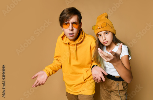 Young teens boy and girl in comfortable clothing, hats and sunglasses standing and expressing misunderstanding