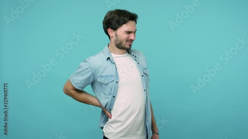 Backache. Tired unhealthy male worker in jeans shirt touching injured sore back, suffering lower lumbar discomfort, muscle pain of overwork, pinched nerve. studio shot isolated on blue background