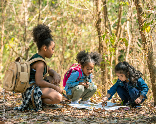 Group of happy pretty little girl hiking together with backpacks and sitting on forest dirt road with looking at the map for exploring the forest. Three kids having fun adventuring in sunny summer day
