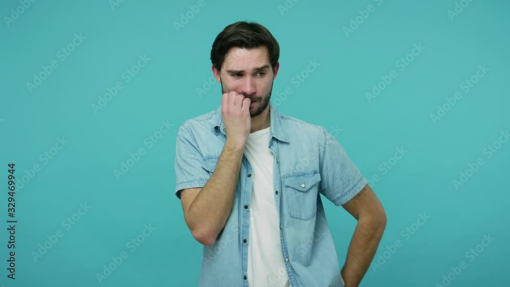 Anxious upset bearded guy in jeans shirt nervously biting nails and fingers, rubbing bristle while thinking over serios troubles, looking scared confused. studio shot isolated on blue background Anxious upset bearded guy in jeans shirt nervously biting nails and fingers, rubbing bristle while thinking over serios troubles, looking scared confused. studio shot isolated on blue background