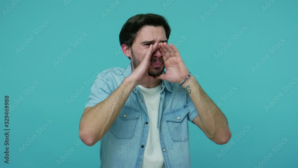 Man shouting advertisement. Young bearded guy in jeans shirt holding arms around mouth and speaking out loud, telling important message, announcement. indoor studio shot isolated on blue background