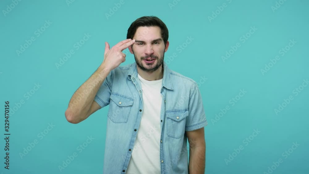 Desperate hopeless bearded guy in jeans shirt pointing finger pistol to head, feeling frustrated showing suicide gesture, shooting himself with hand gun. indoor studio shot isolated on blue background Desperate hopeless bearded guy in jeans shirt pointing finger pistol to head, feeling frustrated showing suicide gesture, shooting himself with hand gun. indoor studio shot isolated on blue background