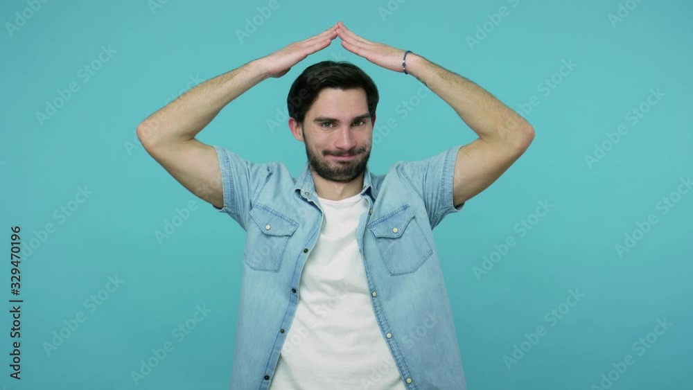 Health and life insurance. Positive young bearded guy in jeans shirt making house roof gesture over head and smiling happily, feeling safe, protected. indoor studio shot isolated on blue background Health and life insurance. Positive young bearded guy in jeans shirt making house roof gesture over head and smiling happily, feeling safe, protected. indoor studio shot isolated on blue background
