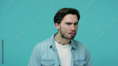 Comical weird bearded guy in jeans shirt watching around making fish face with pout lips, frowning and looking with humorous wary suspicious expression. indoor studio shot isolated on blue background