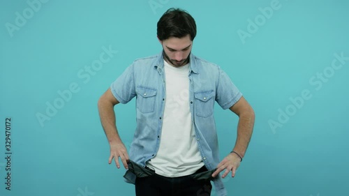Poor student, jobless bearded guy in jeans shirt worried of overspend, showing empty pants pockets having no money, concept of poverty bankruptcy debts. indoor studio shot isolated on blue background