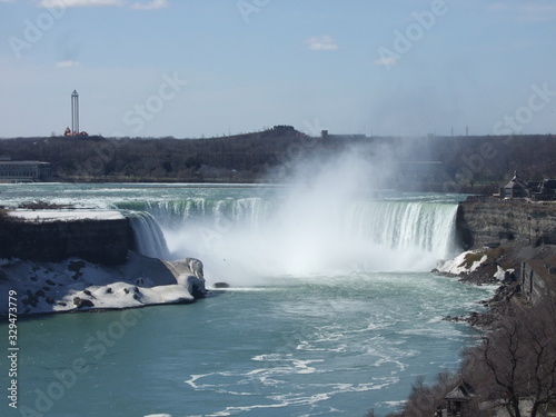 The lovely and lively Niagara Falls with clean, blue water runs down the fall into the river causing a splash over white rocks and boulders and creating a mist in front of the fall with trees behind.