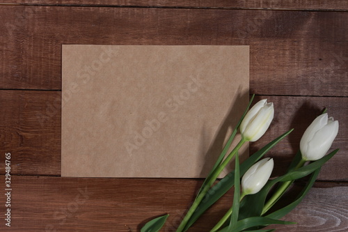 Fototapeta Naklejka Na Ścianę i Meble -  White tulips and blank sheet of craft paper for message or text on brown wooden table. Spring concept. 8 march, women or mothers day, flowers