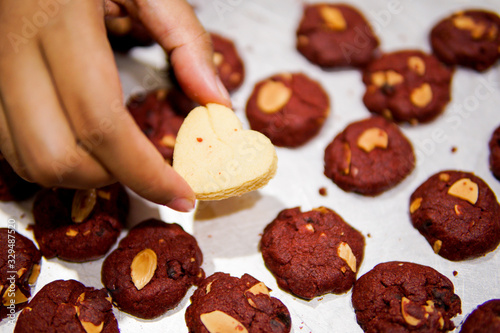 A hand hold a love shape cookies in the middle of red velvet cookies on a tray