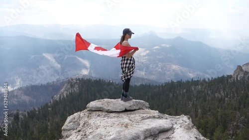 Canadian Girl Cloaked With National Flag Slowmotion. Young Female Standing on Rock Viewpoint With Stunning Mountain Overview photo